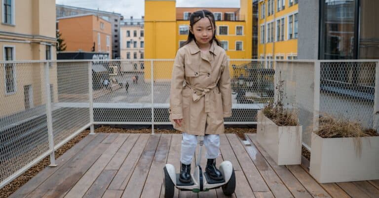 Ein junges Mädchen in einem beigen Trenchcoat und weißen Hosen fährt auf einem weißen Hoverboard auf einem Holzdeck mit Gebäuden im Hintergrund.